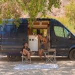A couple enjoying their travel lifestyle, relaxing by a parked camper van in a sunny outdoor setting.