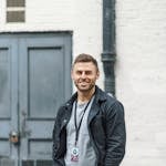 A relaxed portrait of a smiling man in casual attire standing outside against an industrial backdrop.