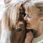 A senior woman lovingly embraces a horse, showcasing warmth and connection in a bright outdoor setting.