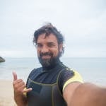 Bearded man in wetsuit taking a selfie at a Portuguese beach, showcasing joy and leisure.
